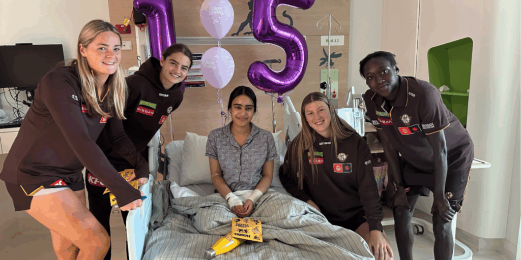 Hawks AFLW players take a photo with a girl in a hospital bed, with ballons behind her that read happy birthday and 15
