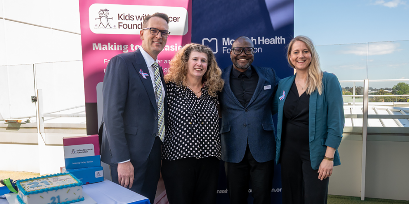 Photo of 4 people standing arm-in-arm in front 2 pull-up banners reading Kids with Cancer Foundation and Monash Health Foundation