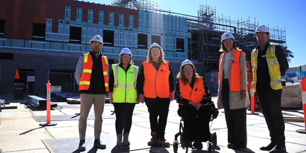 6 people in hi-vis vests and hard hats stand in front of the partially constructed Pakenham Community Hospital