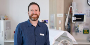 Associate Professor Calum Roberts smiling directly at camera in front of hospital bed