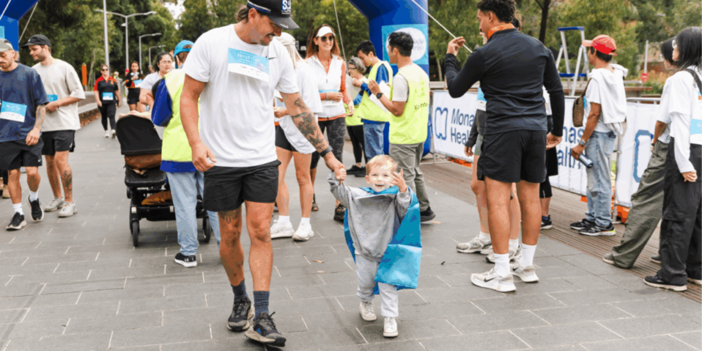 A person holding the hand of a child wearing a blue cape at an outdoor race event.