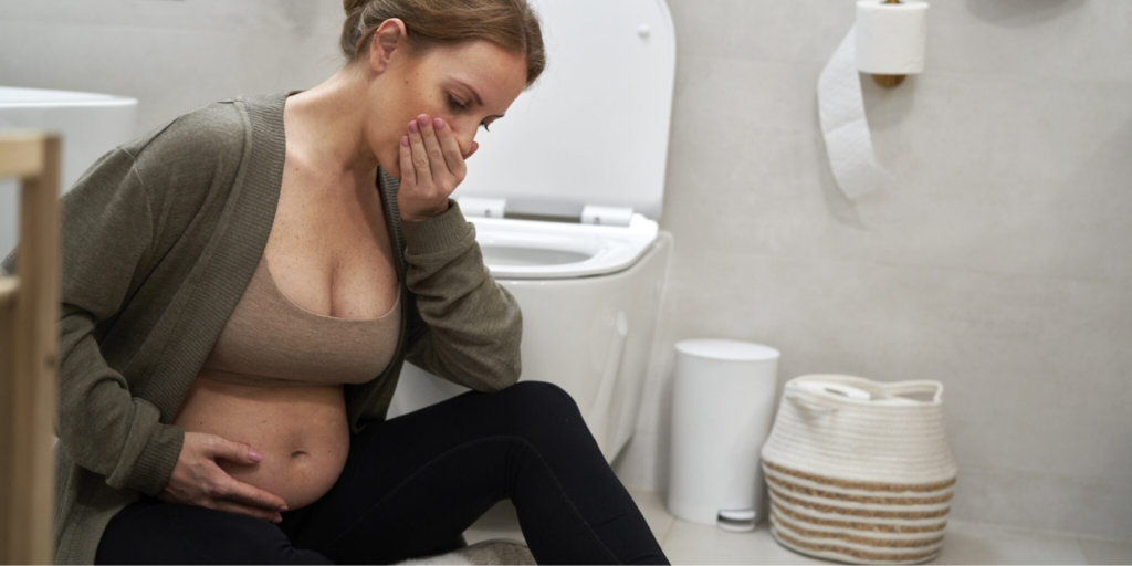 A woman sits on a bathroom floor, holding her stomach and covering their mouth, in front of a toilet.