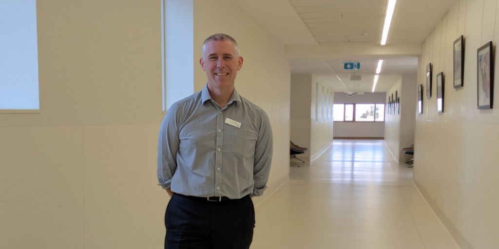 Photo of Simon Craig with grey hair, wearing a grey shirt, employee name badge and black trousers while standing in a hallway