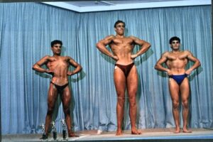 Three male bodybuilders flex their muscles on a podium, photo from another era.