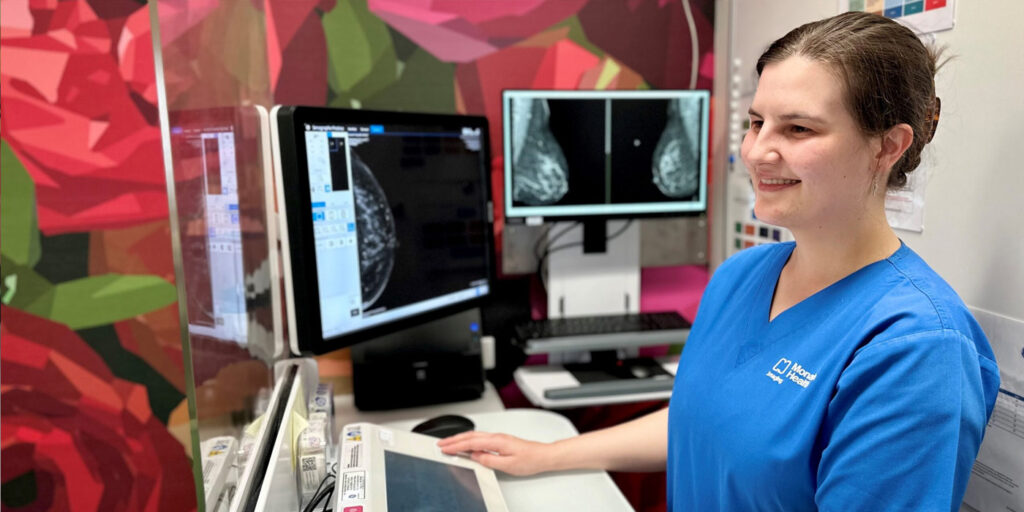 A woman wearing blue scrubs stands in front of a computer screen displaying the results of a Contrast Enhanced Mammography scan