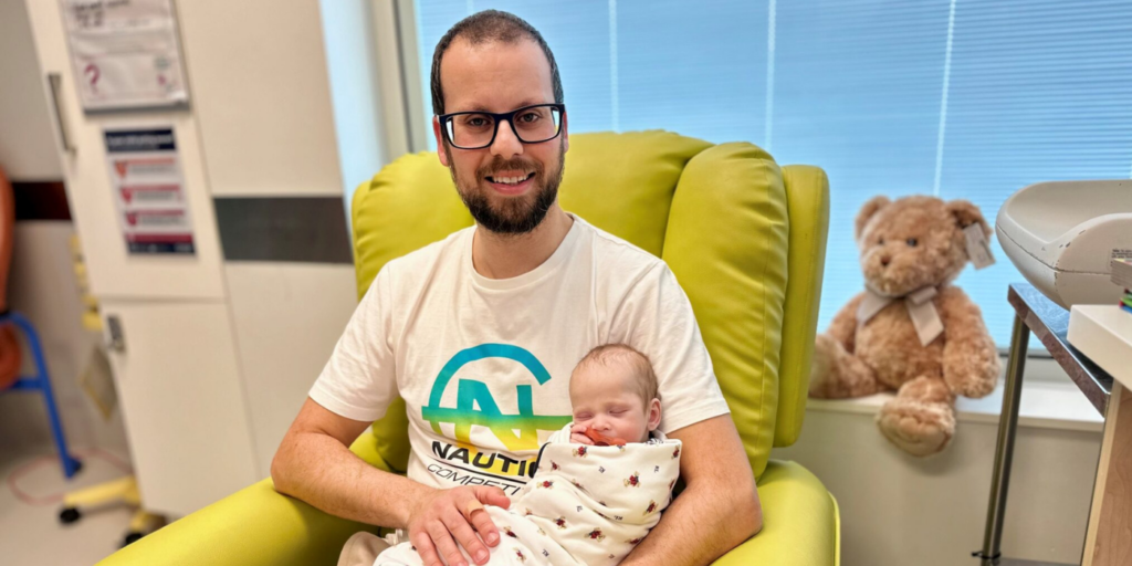 A man sits in a lime green chair holding a baby wrapped in a patterned blanket; a teddy bear sits on a windowsill in the background.