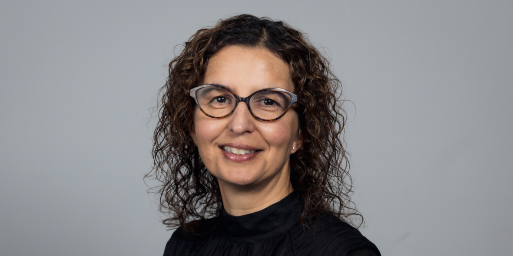 A portrait of Georgia, she has curly brown hair and glasses, smiling against a gray background.