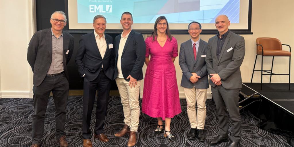 A group of six people stand in a line in front of a projection screen at the Monash Health Foundation Business Breakfast
