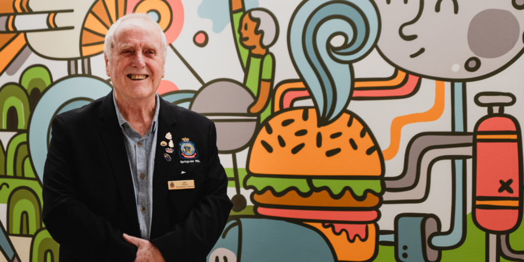 A person in a blazer smiles in front of a colorful mural with cartoon-style images of a burger and other objects