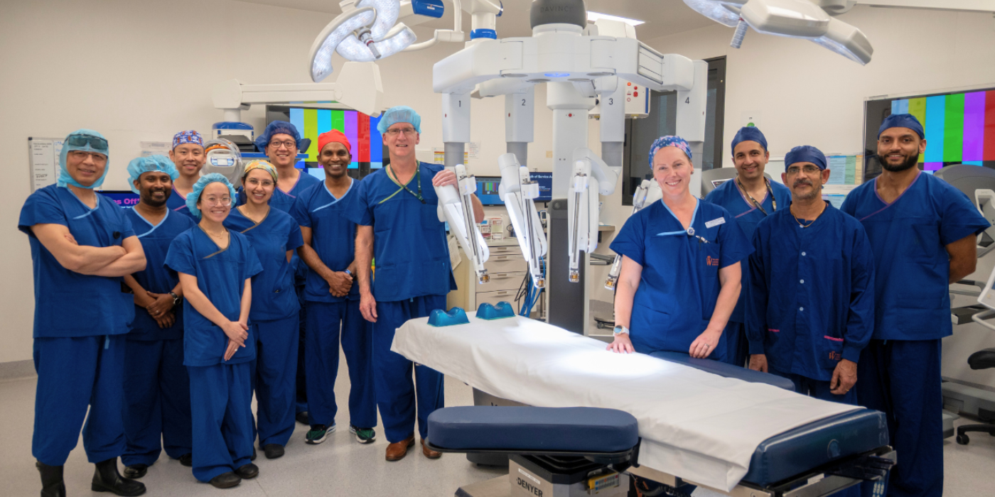 A group of medical professionals wearing scrubs stand in a modern operating room with a surgical robot and an operating table