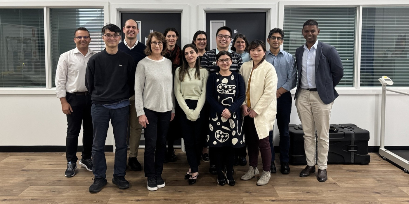 A group of people are standing indoors in front of a white wall with two doors and two windows.