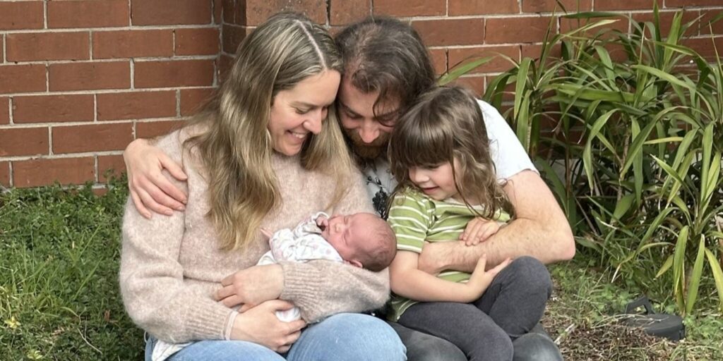 A woman cradling a baby in her arms. Her husband leans in close with his arm around her. Both look adoringly at the baby. A 4-year-old boy cuddles into them.