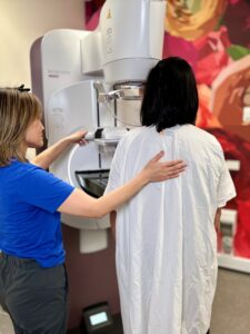 A woman wearing a hospital gown has her back to the camera as she undergoes a mammogram