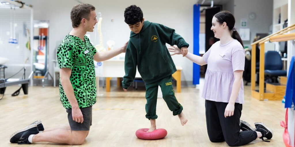 Two allied health workers help a child to balance on one leg