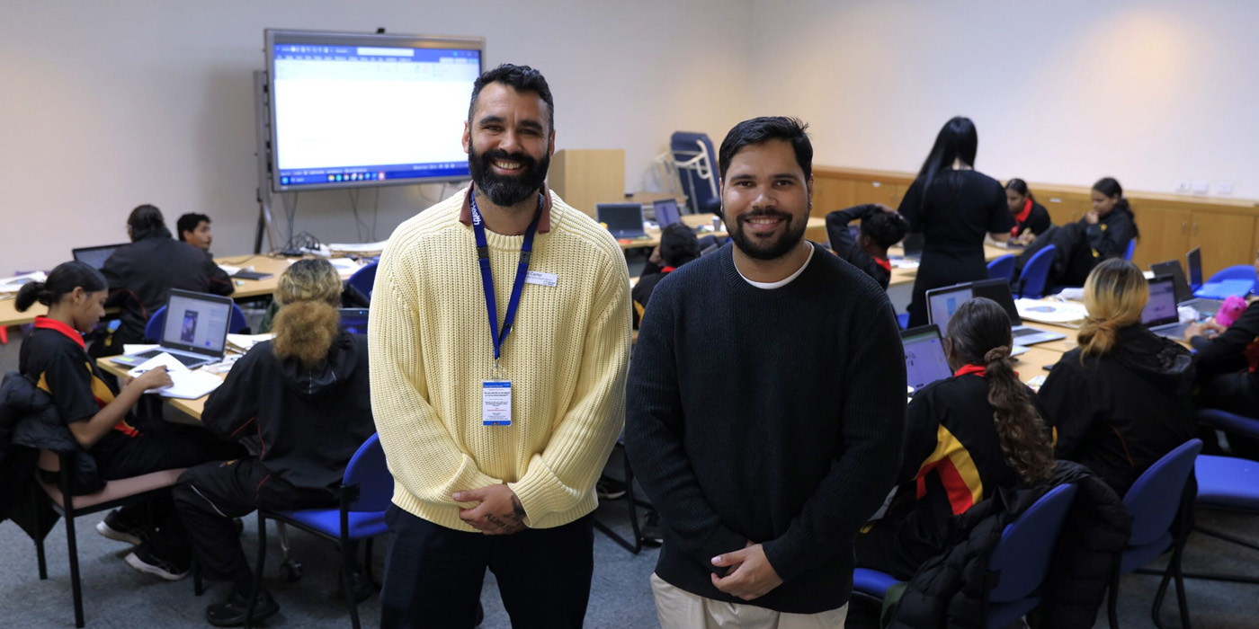 Two men standing in the foreground with a classroom of students on laptops in the background