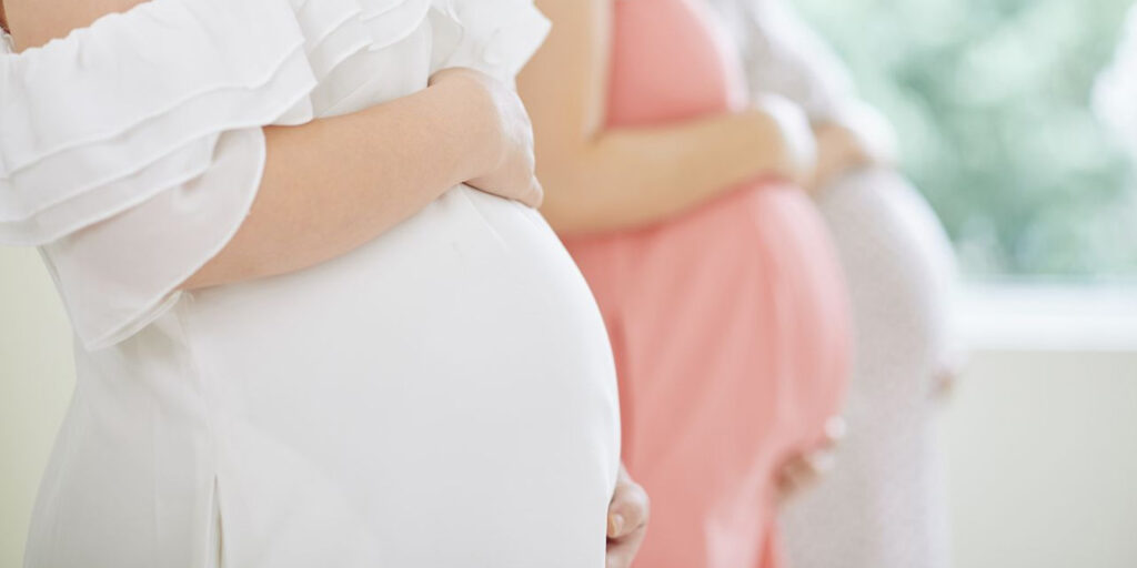 Close-up of a pregnant woman holding her stomach, wearing a white dress, with 2 women in similar poses out of focus in the background
