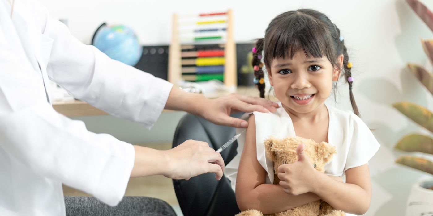 Child holding a teddy bear soft toy receiving a vaccination on her arm from a doctor