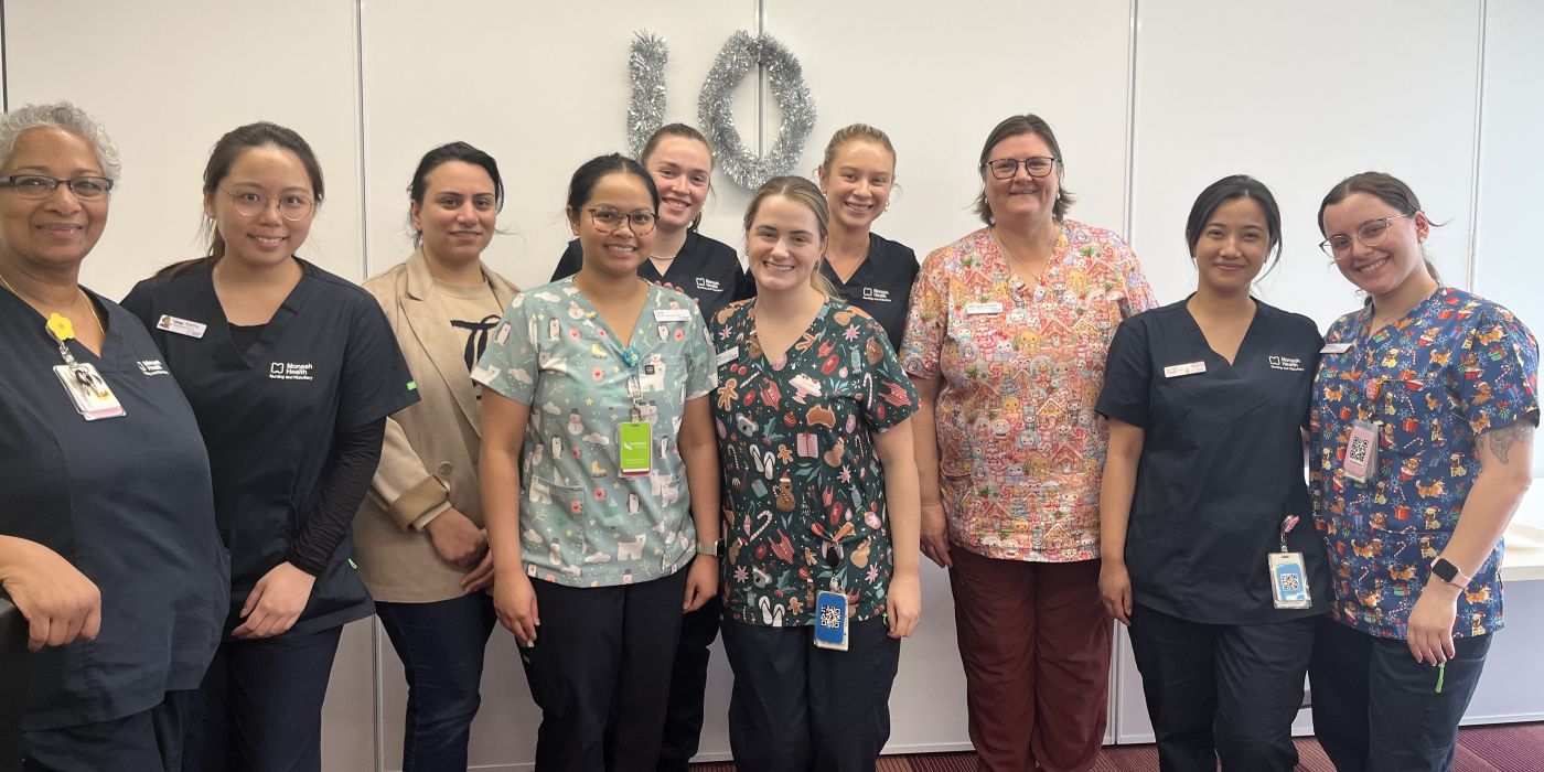 Group of healthcare workers standing together in uniform in front of a white wall with a decorative wreath shaped the number 10.