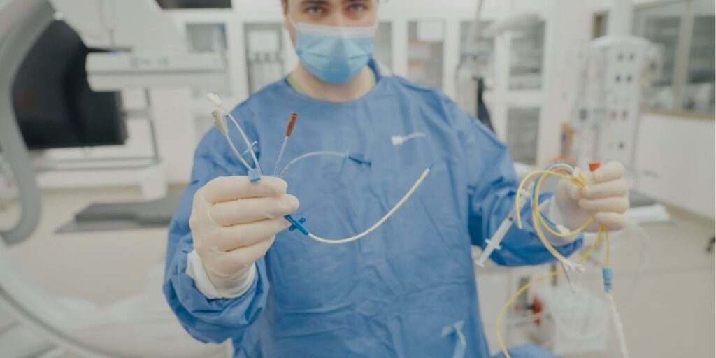 Dr Luke Perry in surgical gown holding various catheters and medical tubing in a clinical setting.