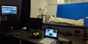 Manikin lying in a hospital bed behind glass, with monitoring equipment and computers in the foreground.