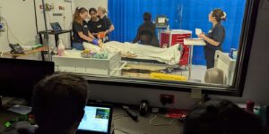 Medical team practices emergency procedures on a mannequin in a simulation room, observed from behind glass.