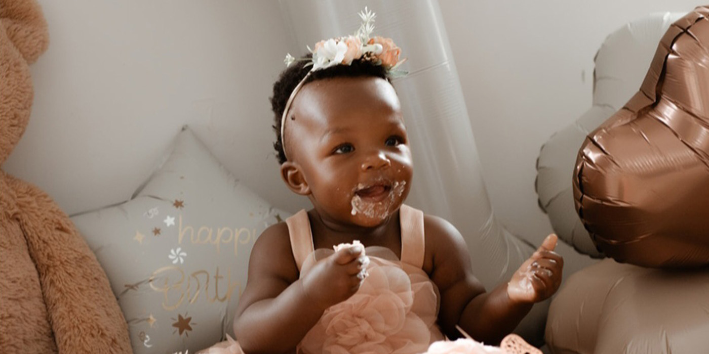 A happy baby sits among balloons with cake in her hand and on her face