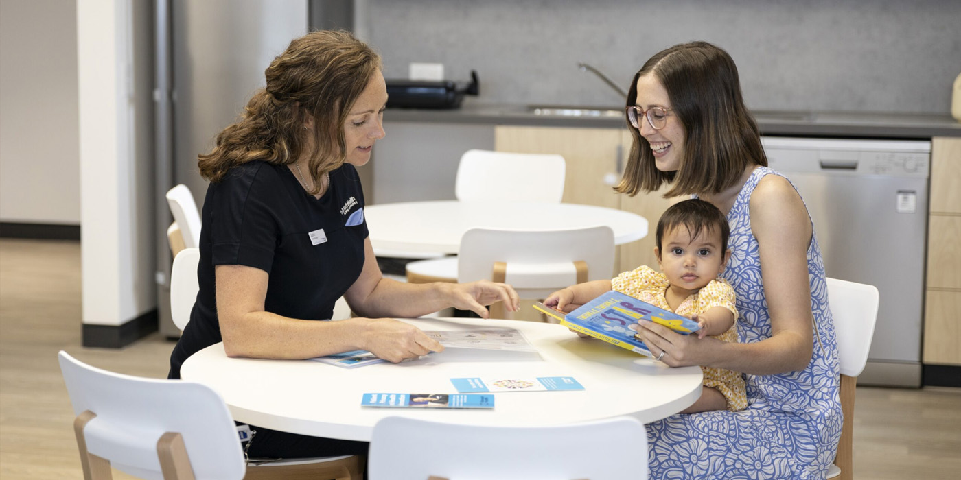 A clinician is reading a brochure to a smiling mother, who has a young child resting on her lap and reading a book