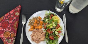 A white plate with flaked tuna, quinoa, roasted sweet potatoes, and mixed salad, and a glass of water with lemon. A napkin and cutlery are laid on the table beside the plate.