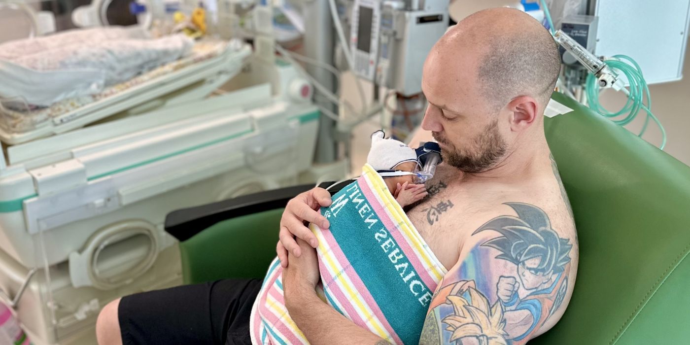 A new father sitting holding his baby daughter to his bare chest. A blanket is wrapped over them.