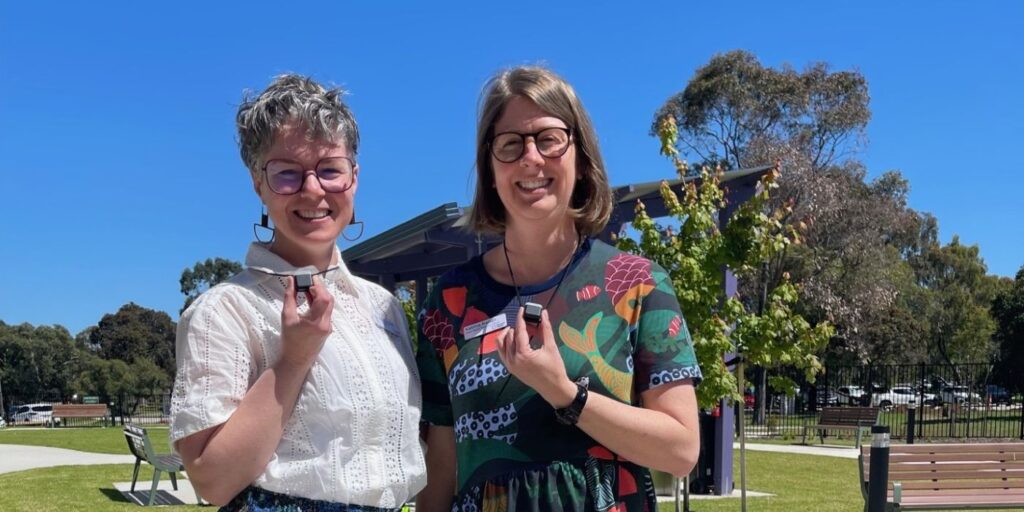 Two people standing outdoors in a sunny park, each holding a small black box the size of a pendant that is laced through a black necklace.