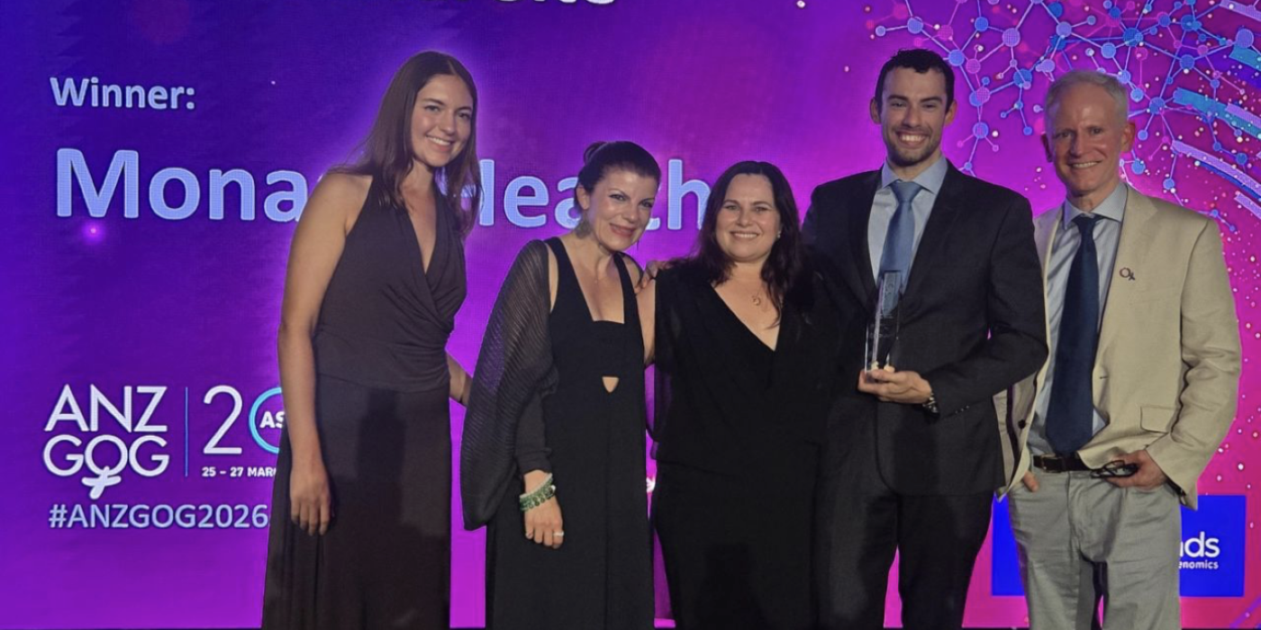 Five formally dressed adults stand on a stage at an awards event, smiling and posing together. One person holds a glass award. Behind them is a large purple screen with text referencing an “Outstanding Clinical Trial Site” and a science-themed graphic, indicating a healthcare or research awards ceremony.