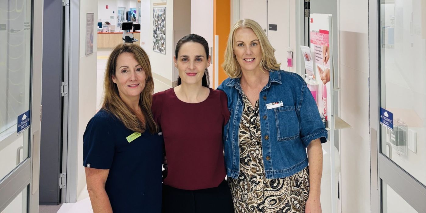 Three women standing together in a hallway, smiling, with office doors and posters in the background.