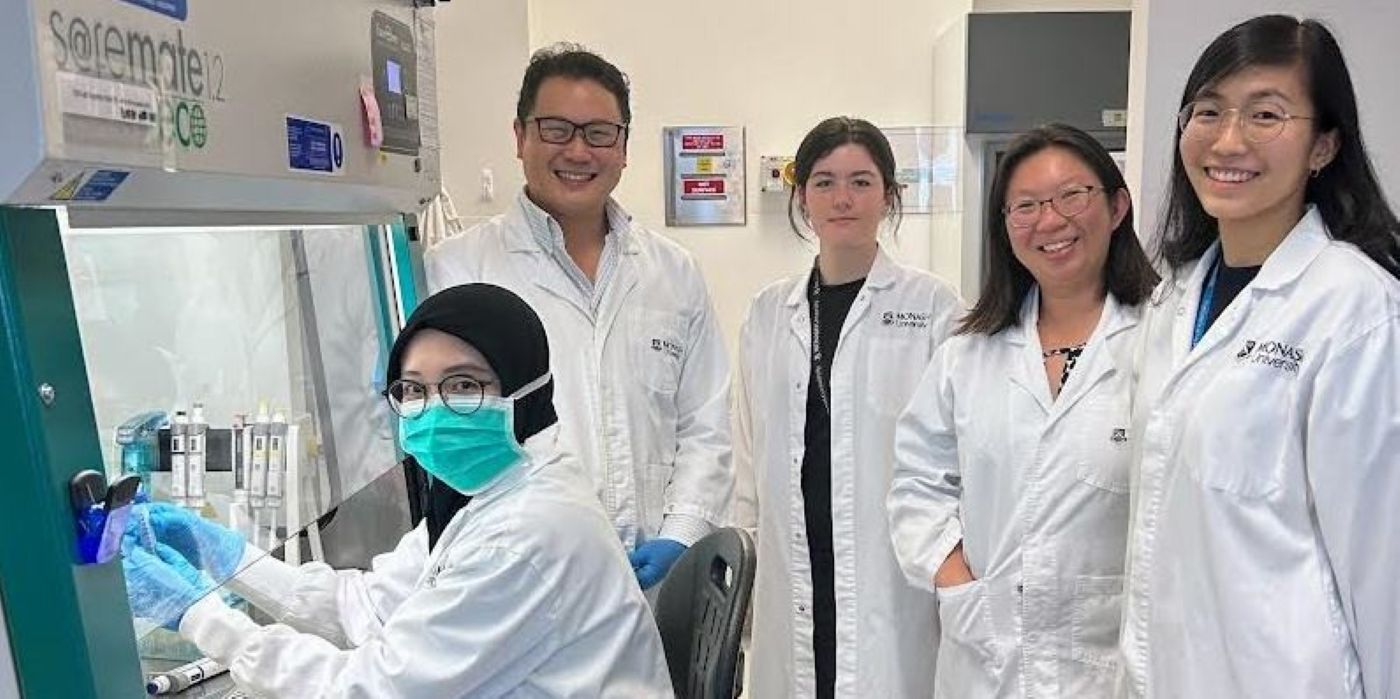 Five people in lab coats, one seated and working at a biosafety cabinet, others standing nearby in a laboratory.