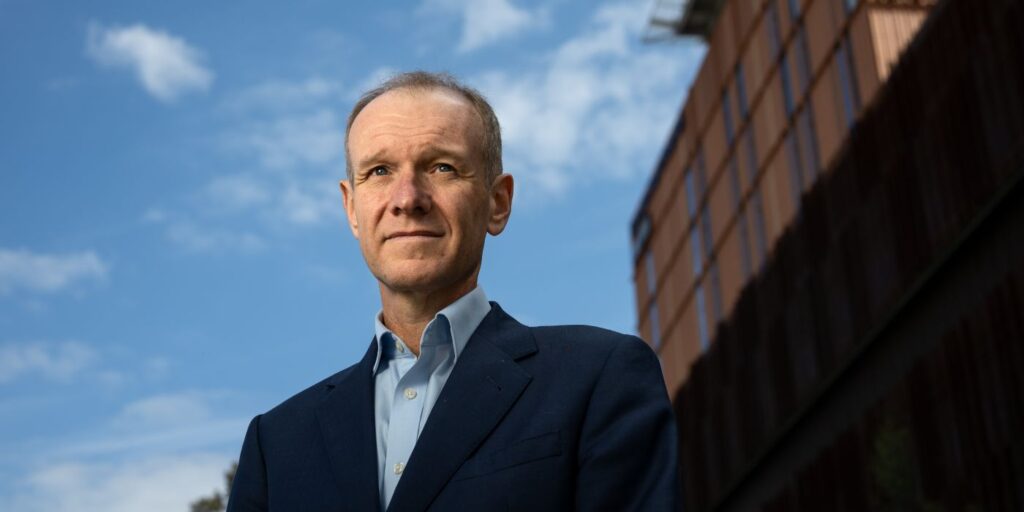 A professional portrait Professor Stephen Nicholls in a navy suit and light blue shirt standing outdoors, photographed from a low angle against a blue sky with scattered clouds. The Victorian Heart Hospital, a modern building with a grid-like facade appears in the background to the right, partially in shadow.