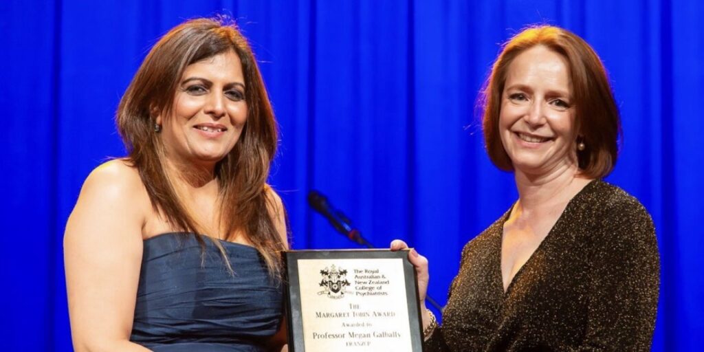 Two women holding an award plaque together on stage with a blue curtain backdrop.