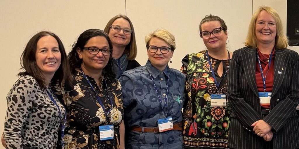 A group of six people standing side by side at an indoor event, facing the camera. Each person is wearing a conference lanyard with a name badge.