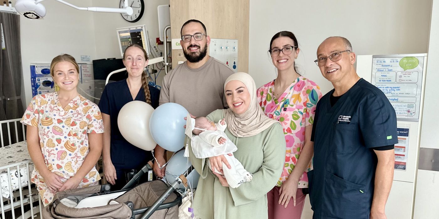 A mother in a headscarf holding her baby son. Her husband stands beside her in a hospital unit. Around them are gathered smiling clinic staff in scrubs.