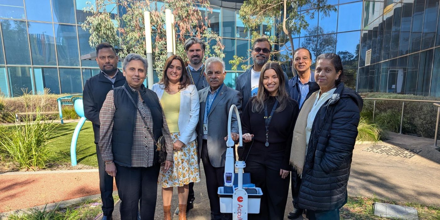Group of nine people standing outdoors in front of Monash Children’s Hospital, gathered around a portable AccuVein vein visualisation device on a wheeled stand.