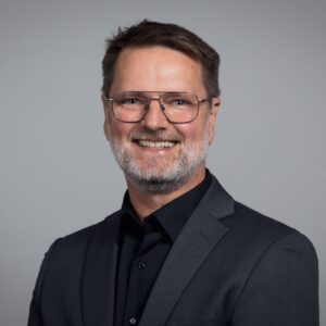 A smiling man wearing glasses, a black shirt, and a suit jacket.