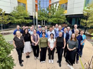 A large group of clinicians and allied health workers standing together outside a modern hospital building, smiling at the camera.