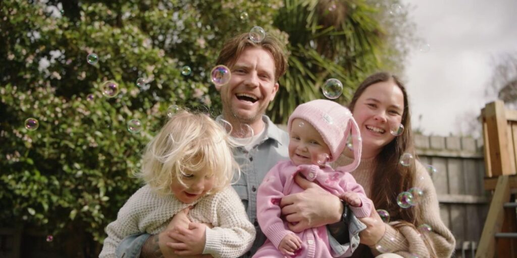 A young family of 4 sitting together outdoors surrounded by bubbles, with greenery and a wooden fence in the background.