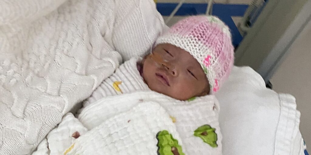 A swaddled premature baby wearing a pink knitted hat is cradled in her mother's arms. The baby has received stem cells from donor umbilical cord blood.