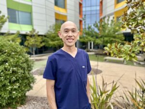 A man wearing blue medical scrubs is standing in a courtyard with a modern hospital building and greenery in the background.