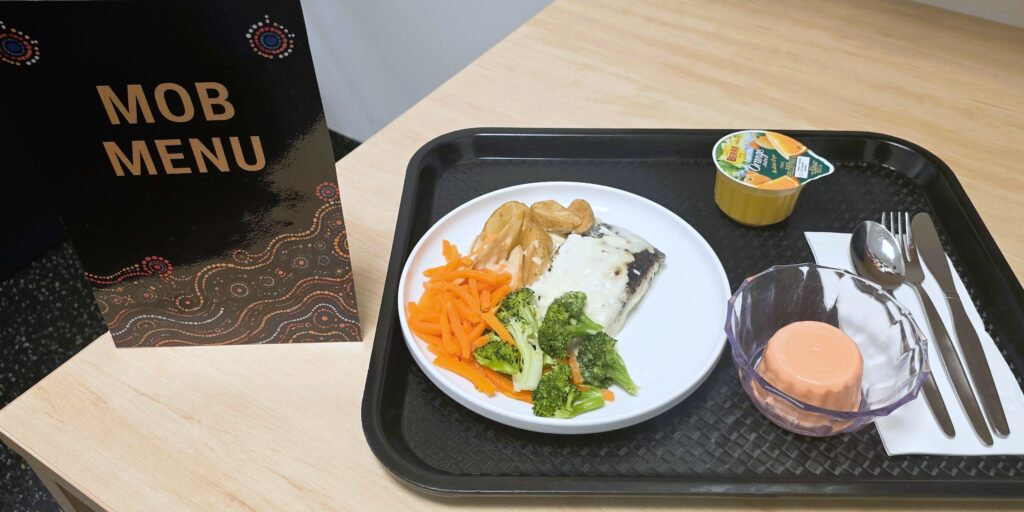 Photo of a tray of food featuring the Kangaroo sausages with steamed vegetables and mashed potatoes, plum Pana cotta and an orange-coloured fruit juice, placed on a table next to the Mob menu sign.