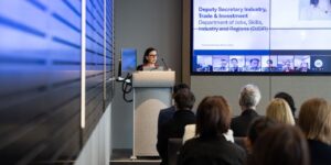Danni Jarrett stands at a lectern addressing an audience in a conference room. A large screen displays a hybrid presentation with virtual attendees joining online, highlighting collaboration and discussion between in-person and remote participants.