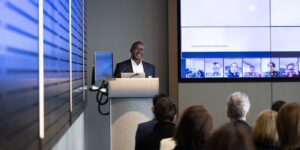 Eugine Yafele speaks from a lectern at the front of a conference room. Attendees are seated facing a large screen showing a hybrid meeting with online participants joining remotely.