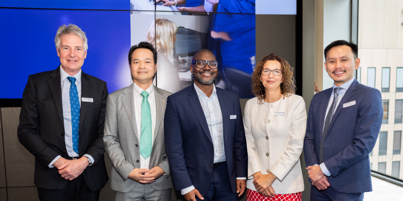 Five people stand together for a group photo at a professional event. The group is positioned in front of a large digital display within a conference venue, with city buildings visible through nearby windows.