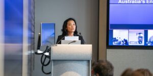 Mimi Tang stands at a lectern delivering a presentation in a conference room. A large screen beside the stage displays presentation slides and virtual participants attending remotely as audience members listen from seated rows.