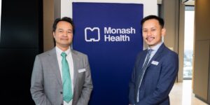 Two people in business attire stand in front of a Monash Health banner at a networking or professional engagement event.